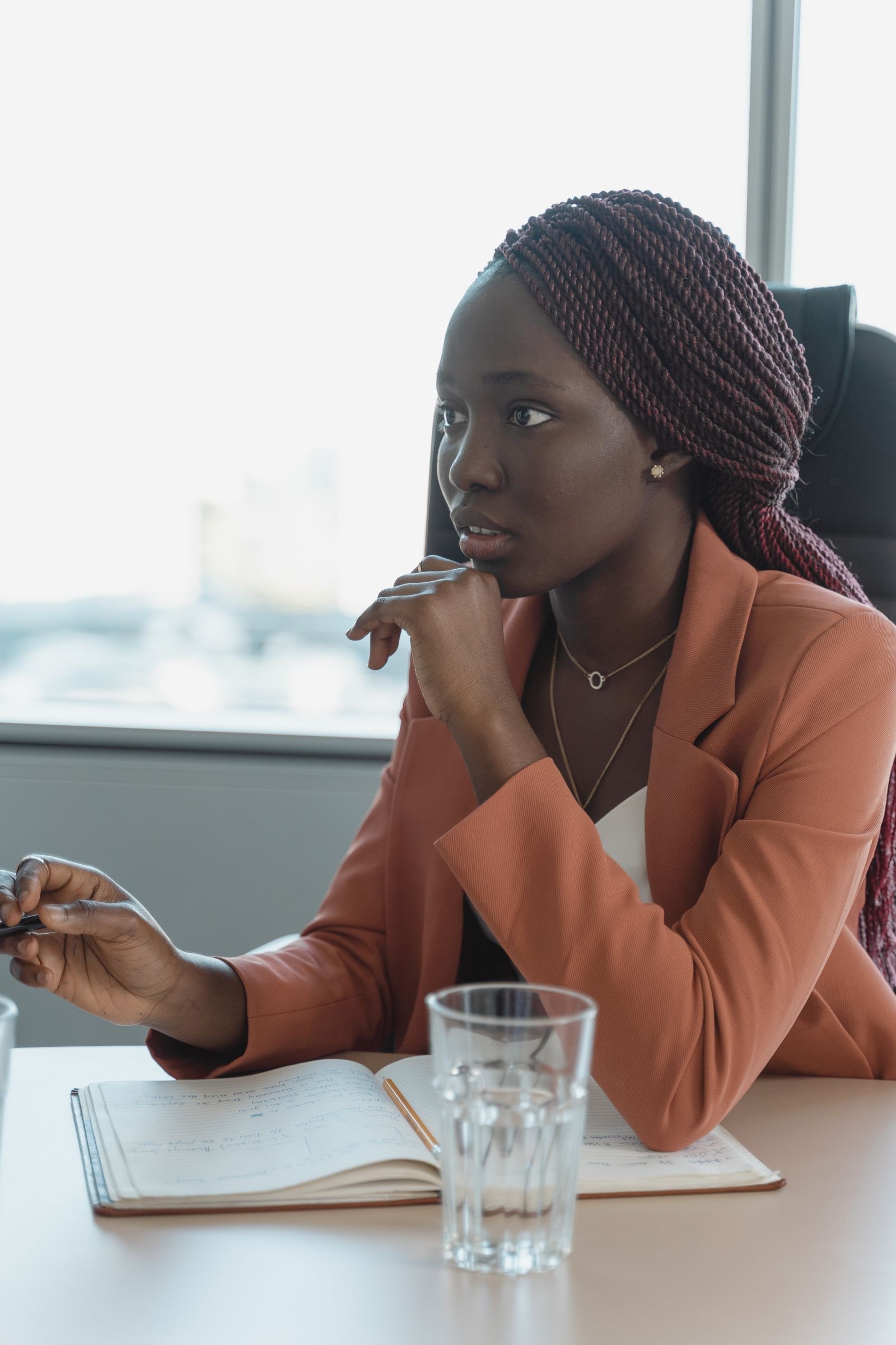 Femme pensive assise à une table de travail, carnet ouvert devant elle, main sous le menton