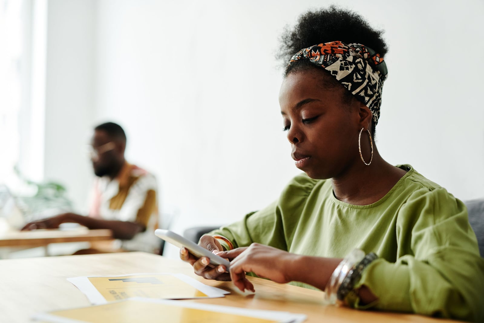 Jeune femme africaine en haut vert et foulard à motifs, en train d'écrire un commentaire sur son téléphone dans un espace de travail lumineux
