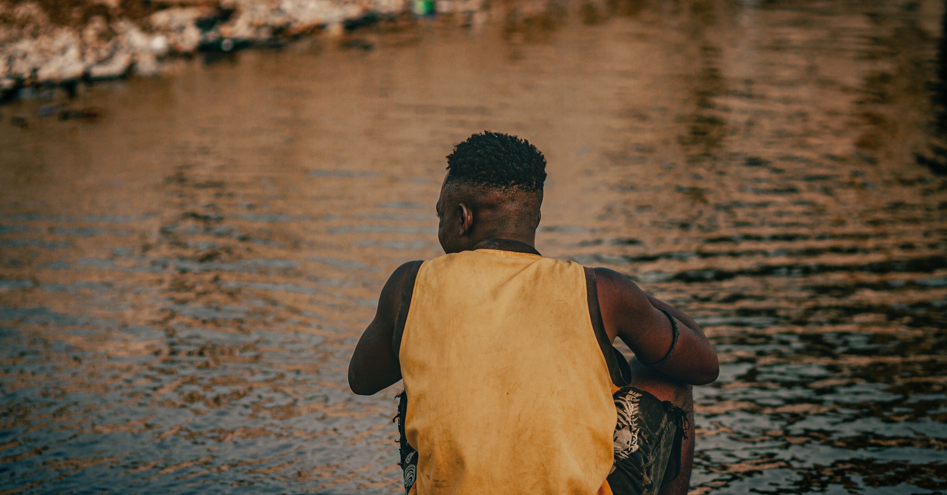 Homme africain assis de dos au bord d'une rivière, seul face à l'eau, lumière chaude du soir