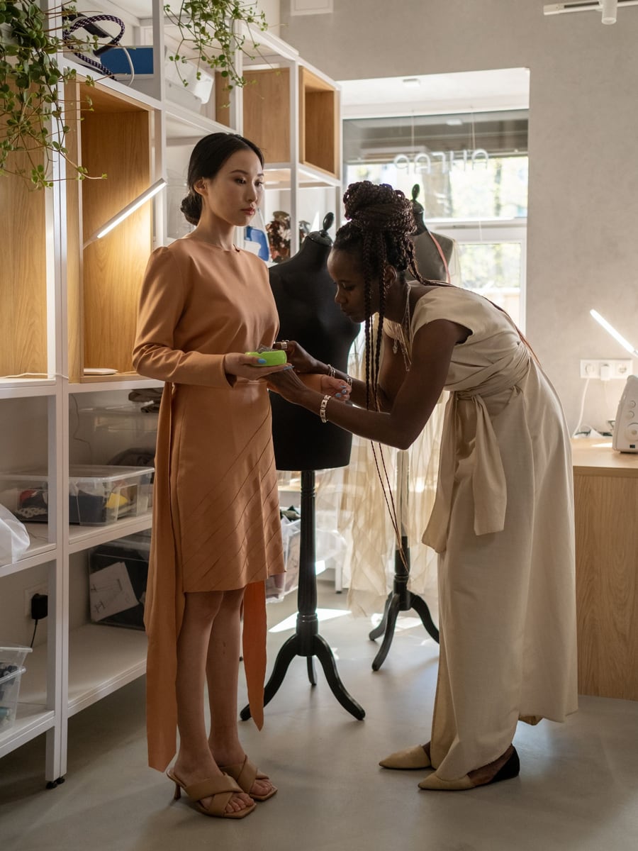 Une créatrice ajuste une robe sur sa cliente debout dans un atelier lumineux