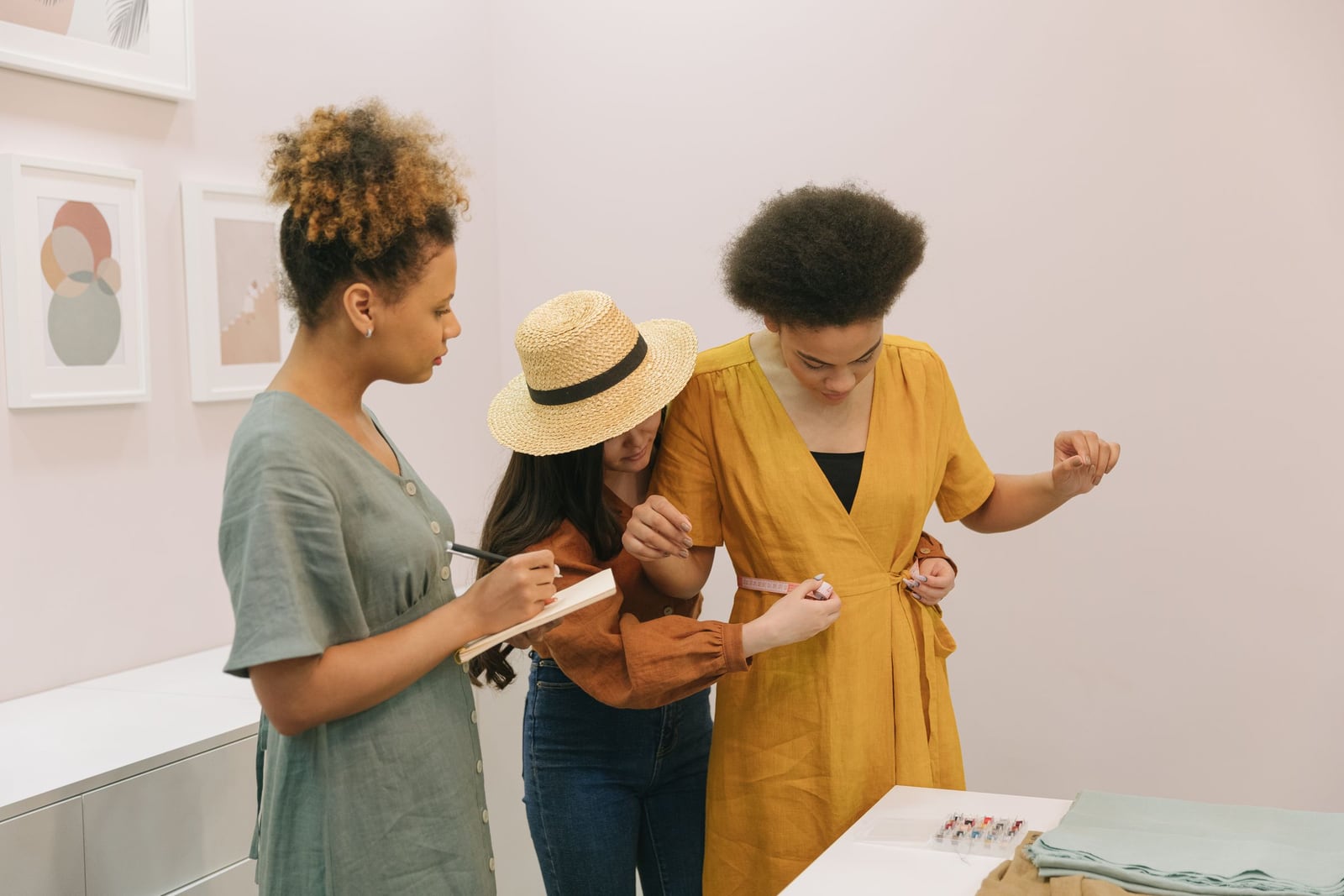 Trois femmes dans un atelier de mode : l'une prend des notes, l'autre mesure à la ceinture, la troisième est en essayage dans une robe ocre