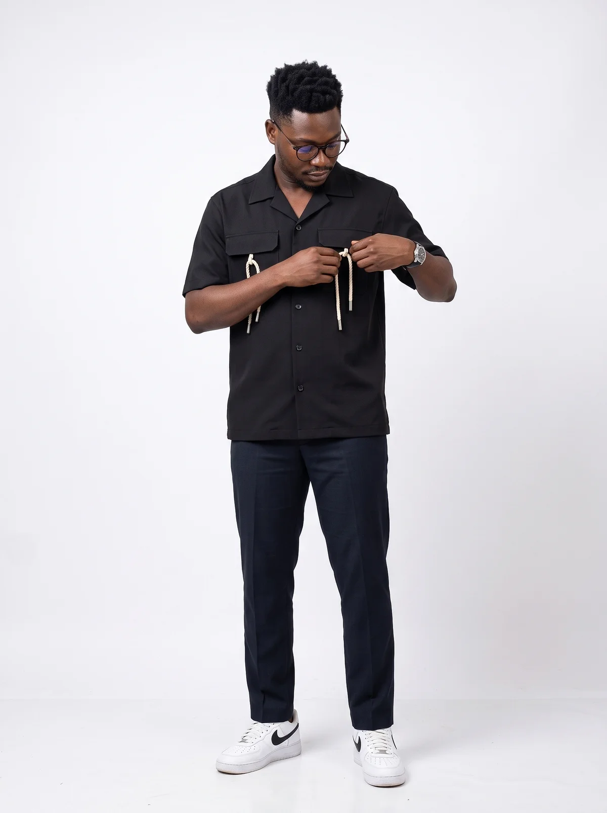 Styliste en chemise noire à cordons et pantalon marine, création Studio a6ko, fond studio blanc